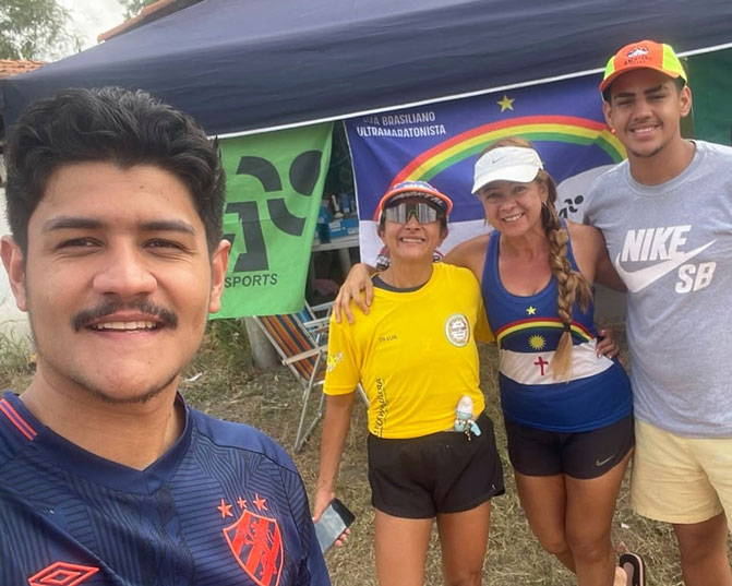 Selfie de quatro pessoas no acampamento: Lua, de camiseta amarela, está ao centro, com uma mulher e dois homens, diante de uma tenda com bandeiras.